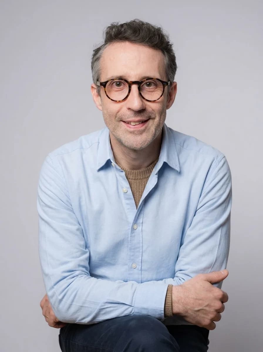 Portrait IA dentiste style corporate chaleureux — chemise bleu clair sur pull col rond beige, posture détendue bras croisés, fond neutre